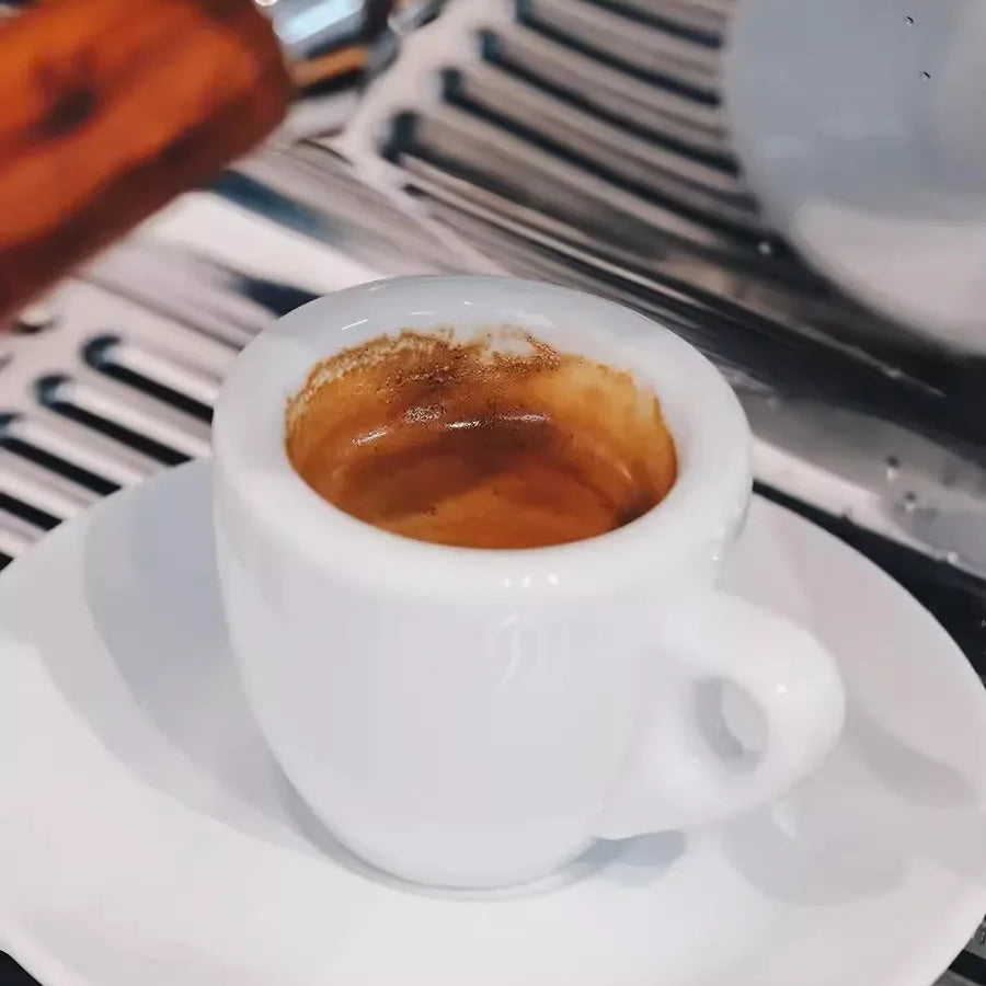 Espresso being poured into a white cup on a saucer with a coffee machine in the background.