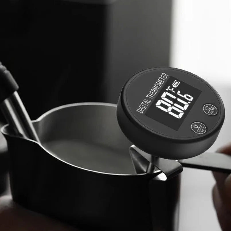 A close-up of a digital thermometer probe attached to a black milk pitcher, with a hand holding the handle and another hand holding the pitcher. The thermometer display reads 80.
