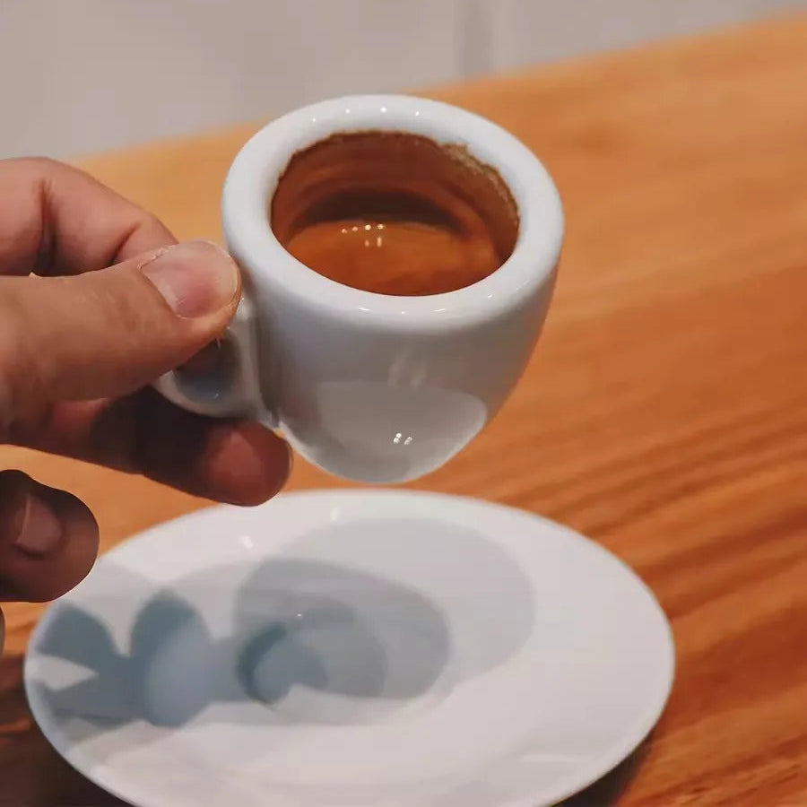 Hand holding a small white cup filled with espresso on a wooden surface.
