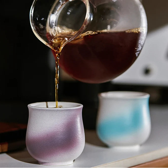 Tea being poured from a glass teapot into a white cup with a blurred background